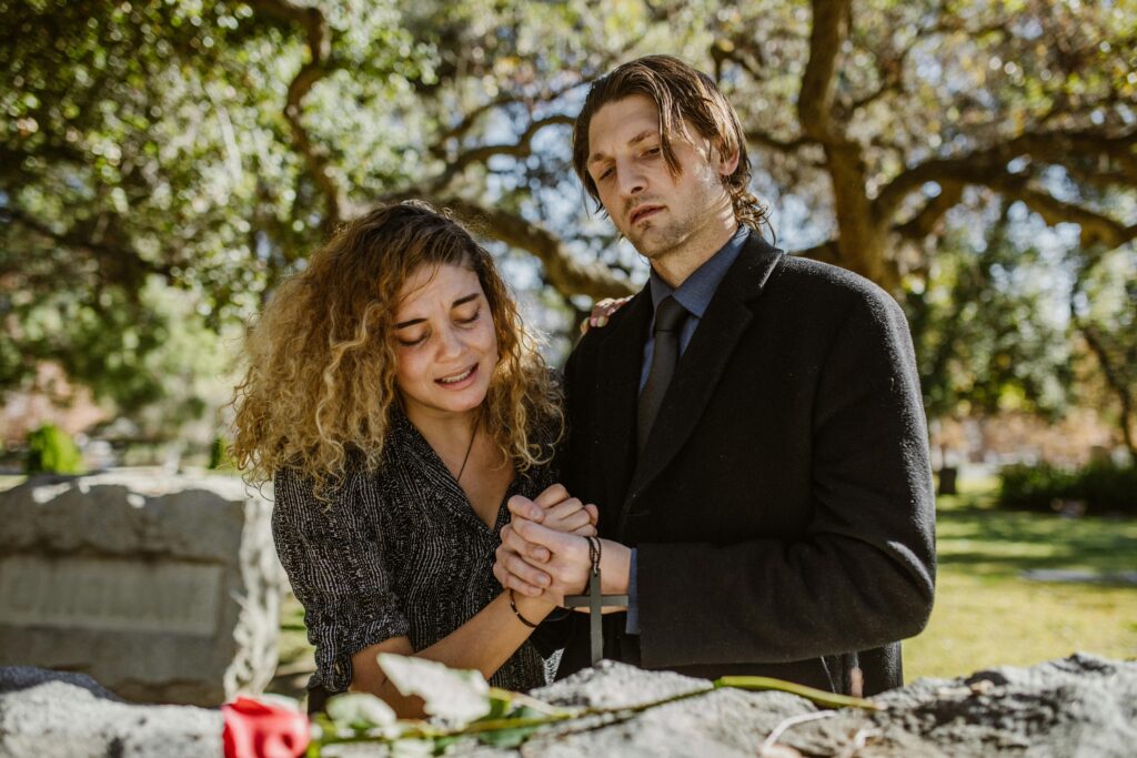 A couple mourns together, holding hands at a grave in a serene cemetery setting.